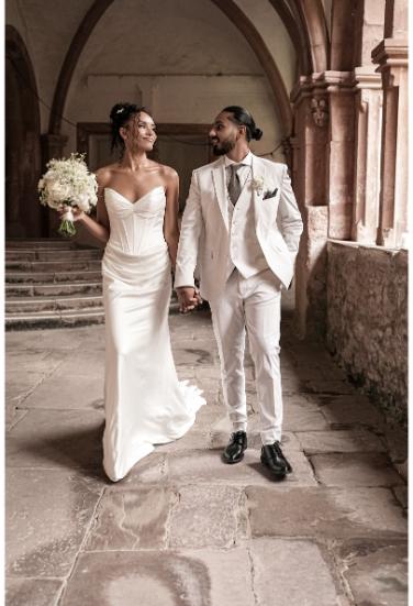 Die Braut und der Bräutigam spazieren Hand in Hand durch die Gänge eines historischen Gebäudes, sie tragen ein stilvolles Hochzeitskleid und einen eleganten Anzug.