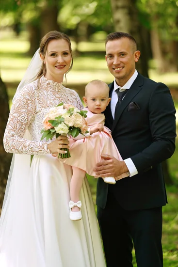 Braut und Bräutigam posieren mit ihrem Kind in einem Park, die Braut im Spitzenkleid mit Blumenstrauß, der Bräutigam im dunklen Anzug – ein emotionales Hochzeitsfoto im Grünen.