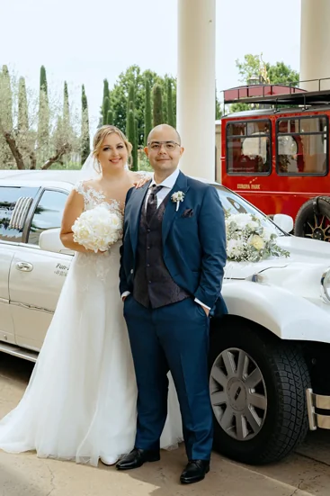 Braut und Bräutigam posieren vor einer weißen Stretchlimousine – die Braut im eleganten Spitzenkleid mit weißem Brautstrauß, der Bräutigam im blauen Hochzeitsanzug mit Weste und Krawatte. Im Hintergrund ein roter Oldtimer-Bus.
